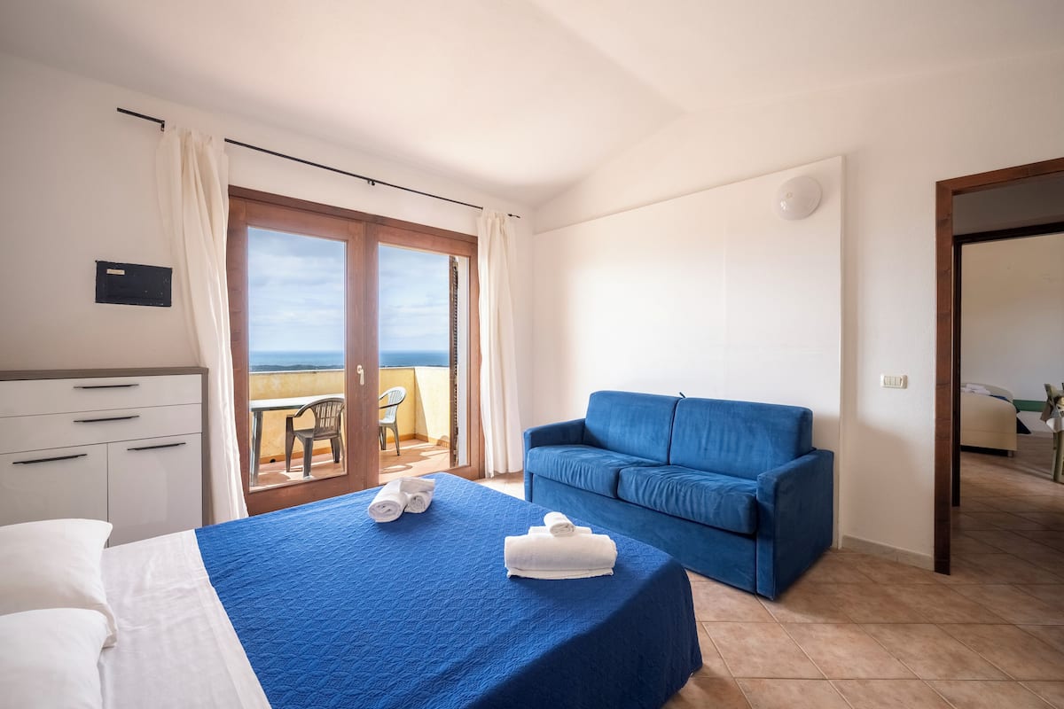 Une chambre d’hôtel avec un lit, un canapé bleu, un balcon avec vue et un bureau avec des tiroirs.
