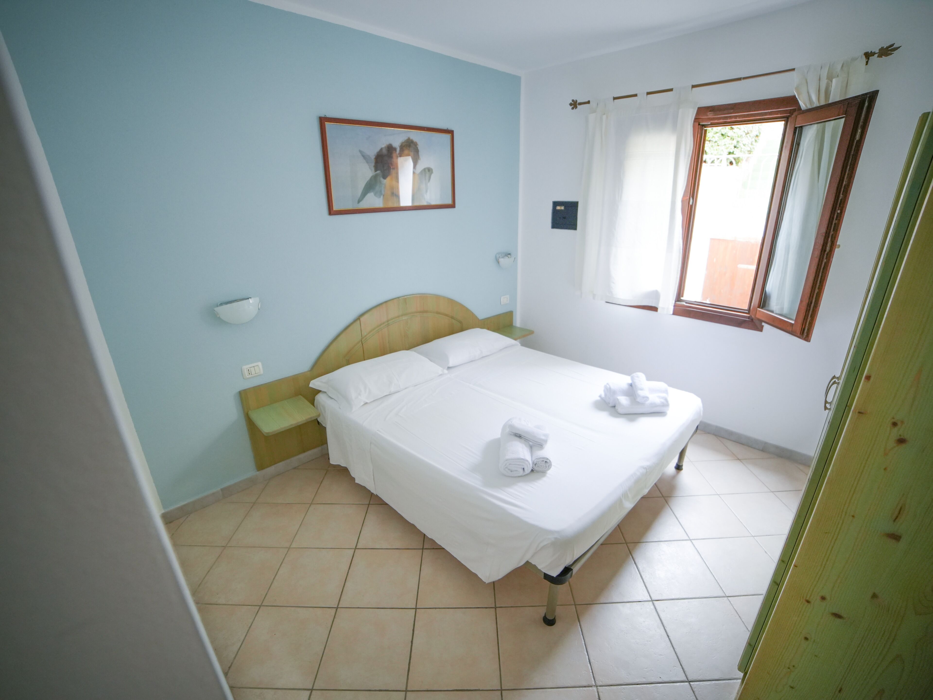 A neatly made bed with white linens and towels, a bedside table, a nightstand, a wall-mounted light, a window with curtains, and a framed picture on the wall.