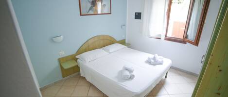 A neatly made bed with white linens and towels, a bedside table, a nightstand, a wall-mounted light, a window with curtains, and a framed picture on the wall.