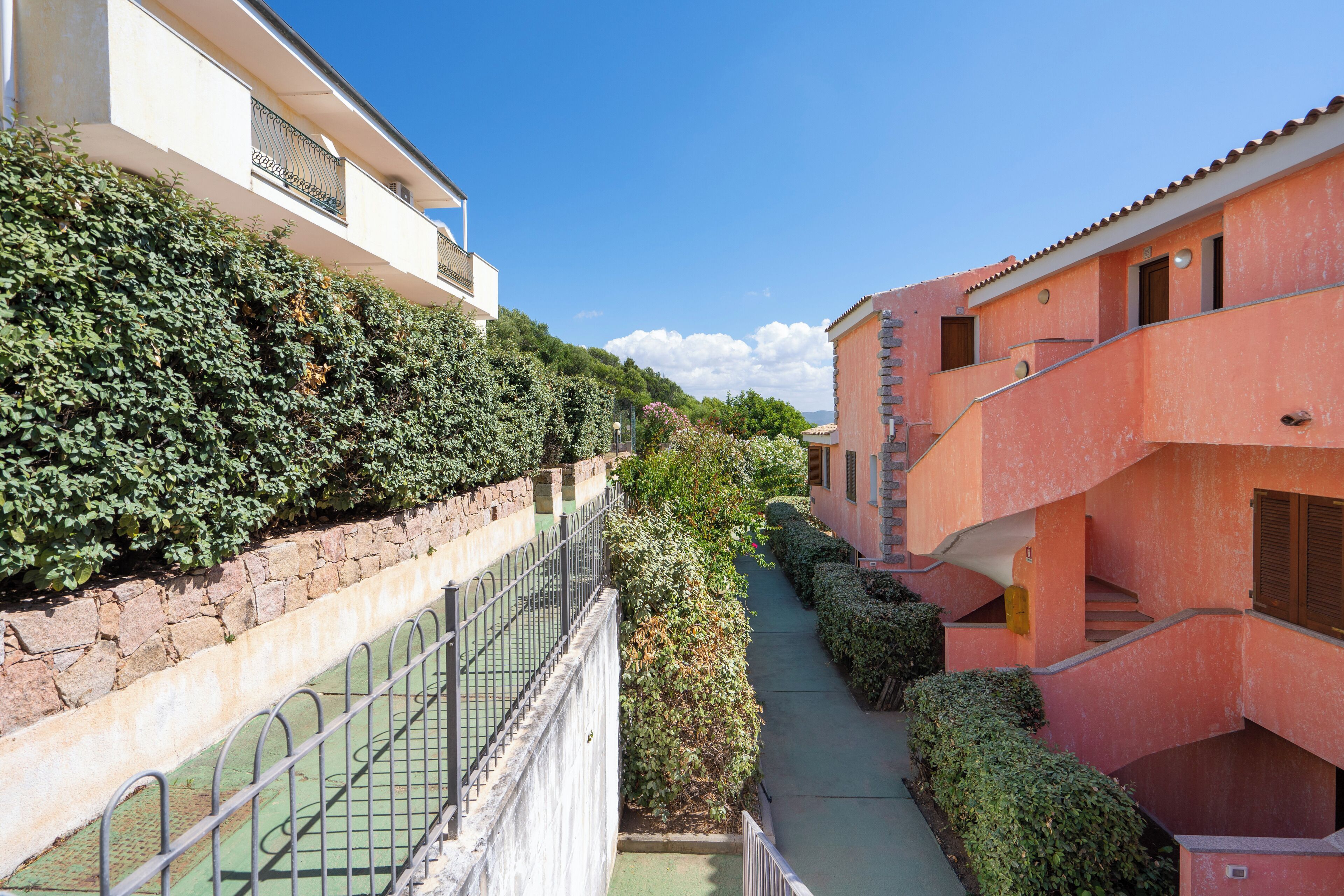 A narrow pathway flanked by buildings with balconies and a stone wall on one side.
