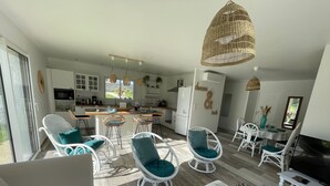 Ein geräumiger Wohnbereich mit einer großen Couch, weißen Korbstühlen, einem Esstisch und einer Küche im Hintergrund.
