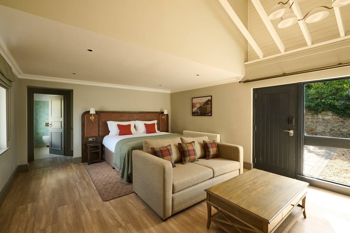 Quarto com cama, sofá, mesa de madeira e uma janela grande com vista para área verde.
