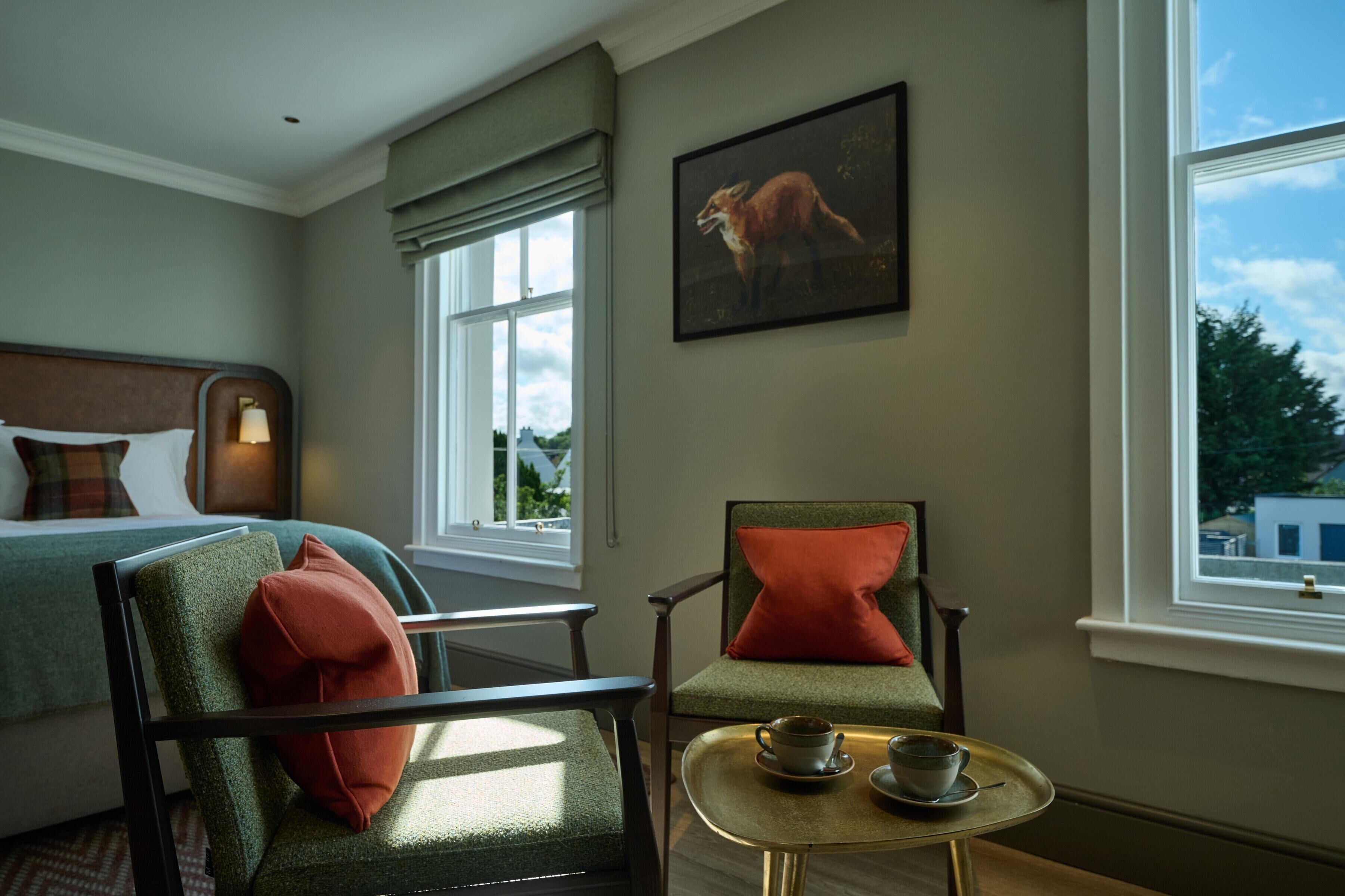 A hotel room with a bed, two chairs, a small table with cups, a painting of a fox, and a window with a view of trees and buildings.