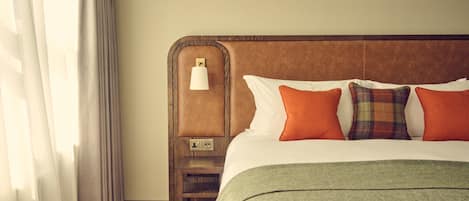 Una cama bien hecha con un edredón verde, dos almohadas naranjas y una almohada a cuadros. Una mesita de noche de madera con una lámpara está al lado de la cama.