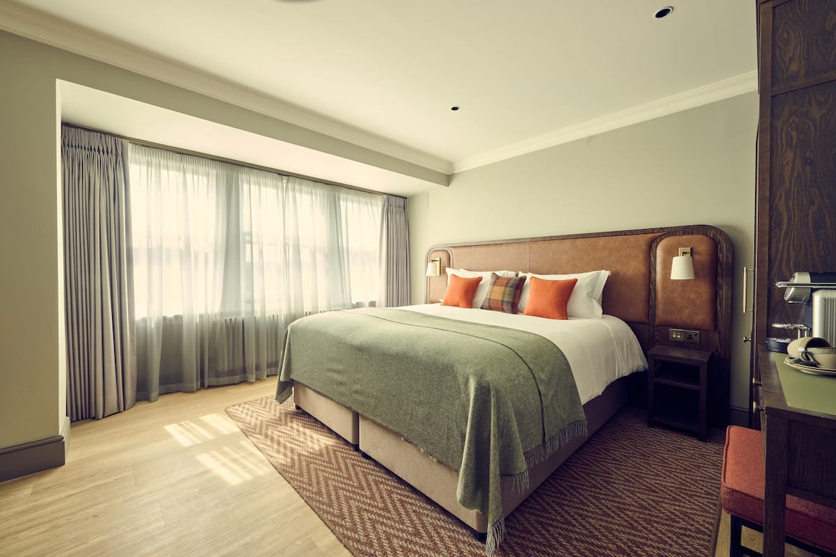 Quarto de hotel com uma cama grande, uma mesa de cabeceira, uma escrivaninha e vista para o exterior através de cortinas translúcidas.