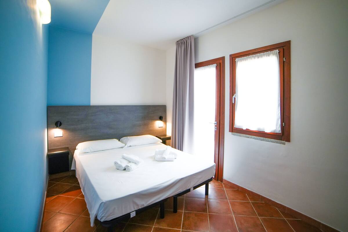 A hotel room with a bed, two bedside lamps, a window with curtains, and a wall-mounted light fixture.