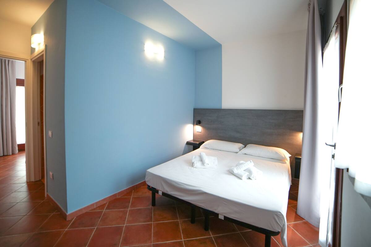 A hotel room with a bed, a nightstand, a wall-mounted lamp, and a window with curtains.