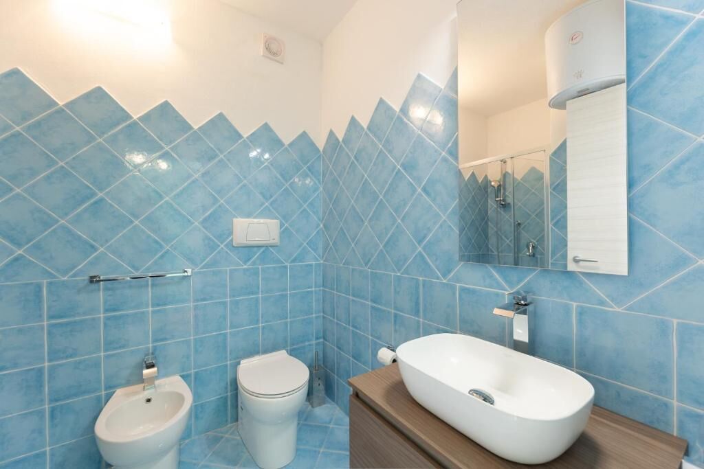 Un baño con azulejos blancos y patrón de diamantes azules, un bidé blanco, un inodoro y un mueble de tocador de madera con un lavabo blanco.