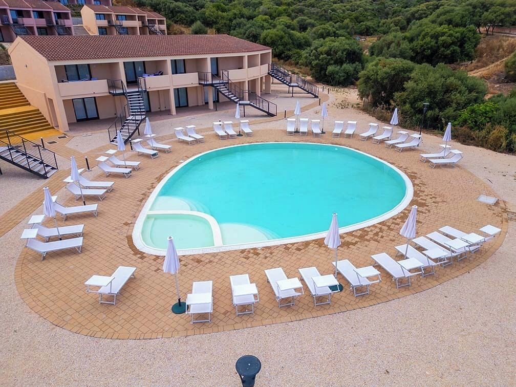 A swimming pool surrounded by sun loungers and umbrellas, with a building in the background.