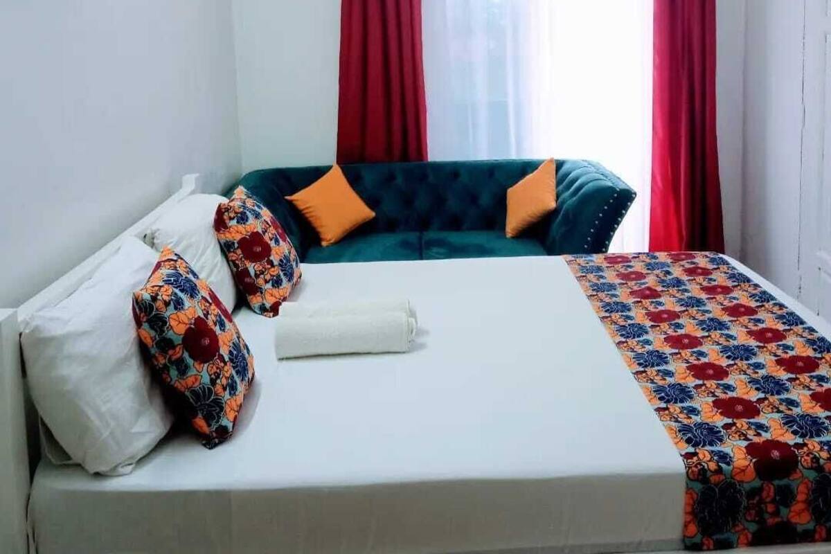 Una cama con sábana blanca, dos almohadas naranjas y un cabecero azul. Hay cortinas rojas en la ventana y una pared blanca con un adorno.