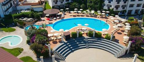 Um resort com uma grande piscina central, espreguiçadeiras e uma escadaria que dá acesso à área da piscina.