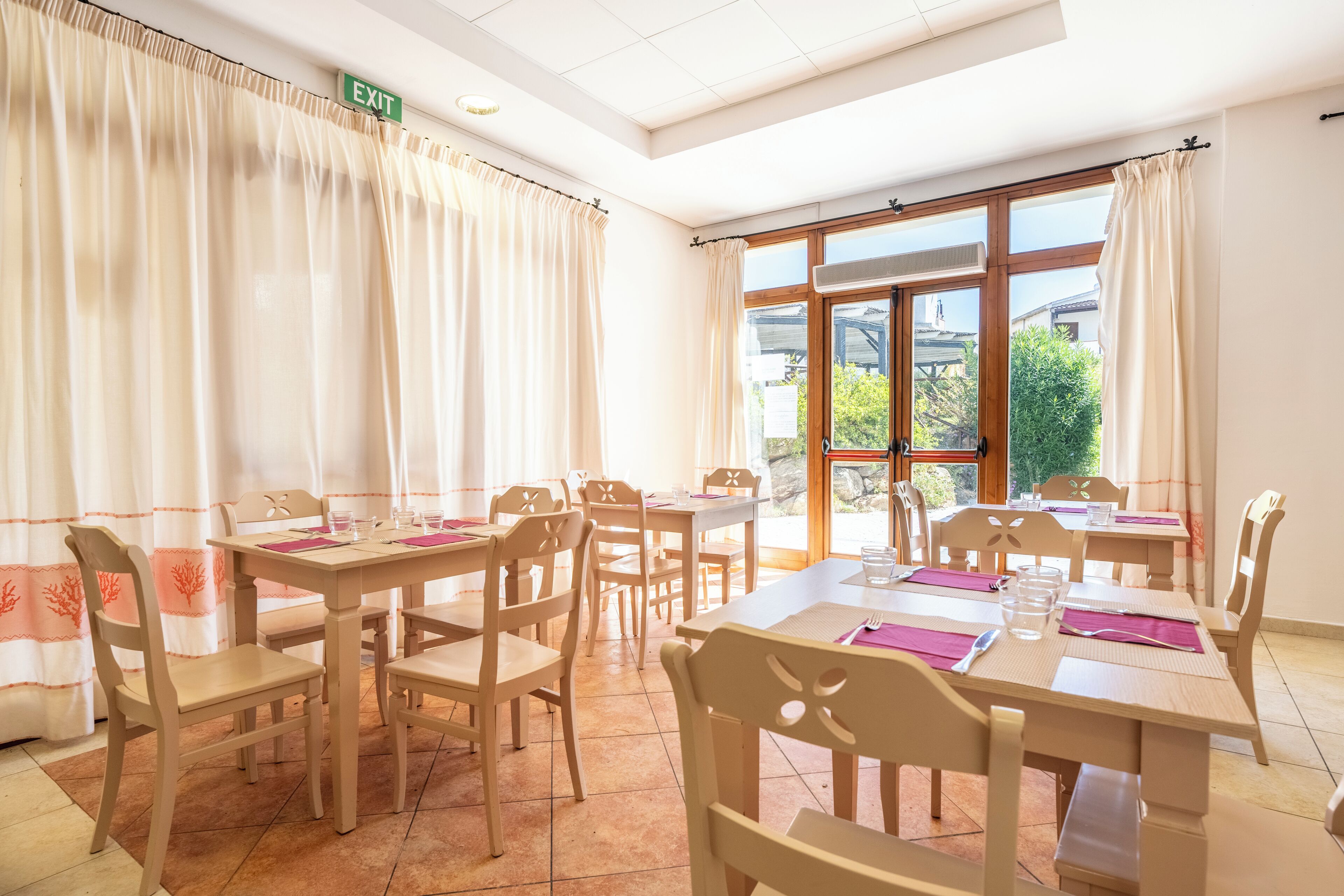 A dining area with wooden tables and chairs, a green exit sign, and large windows with curtains.