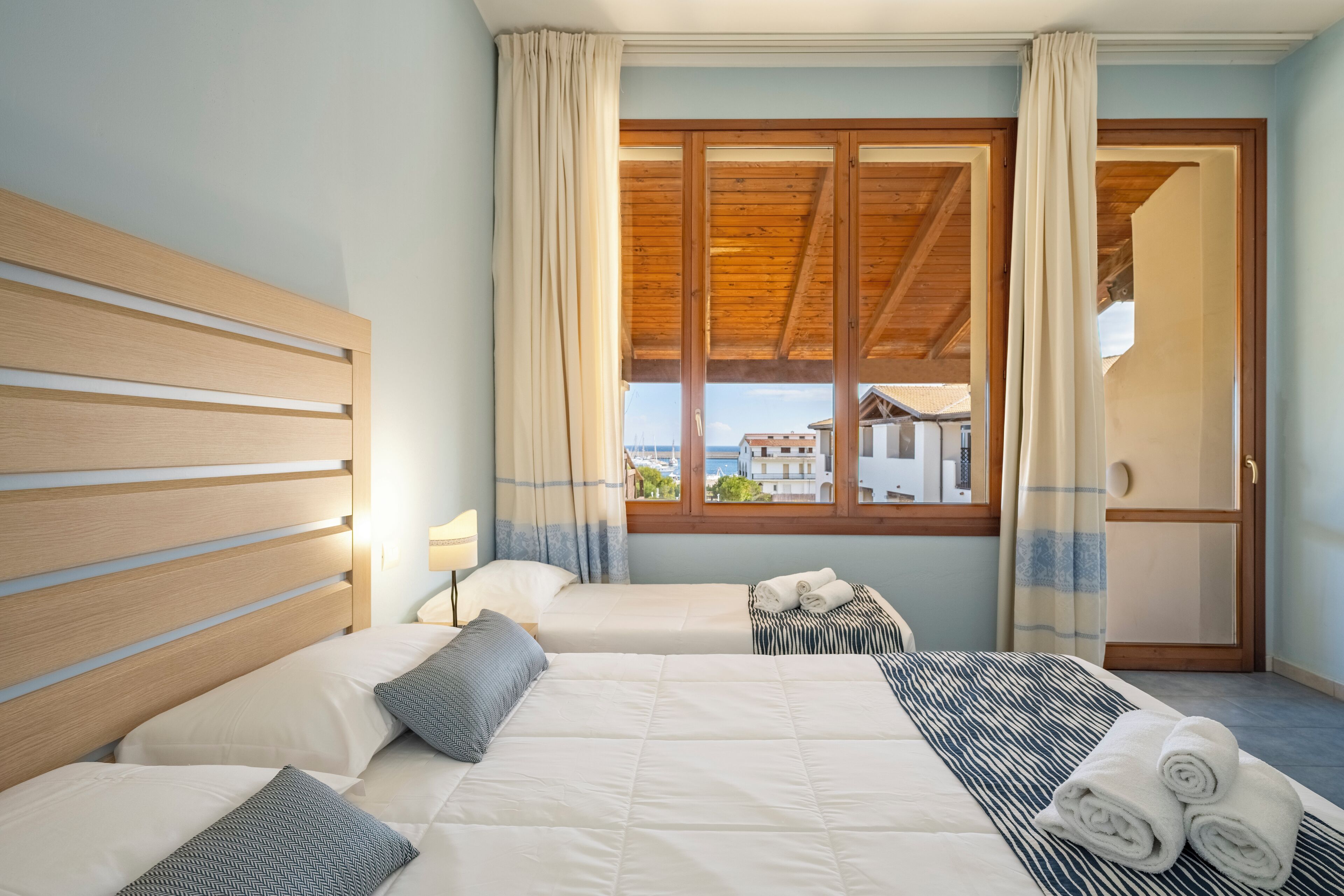A hotel room with two beds, a wooden headboard, a large window with curtains, and a view of buildings outside.