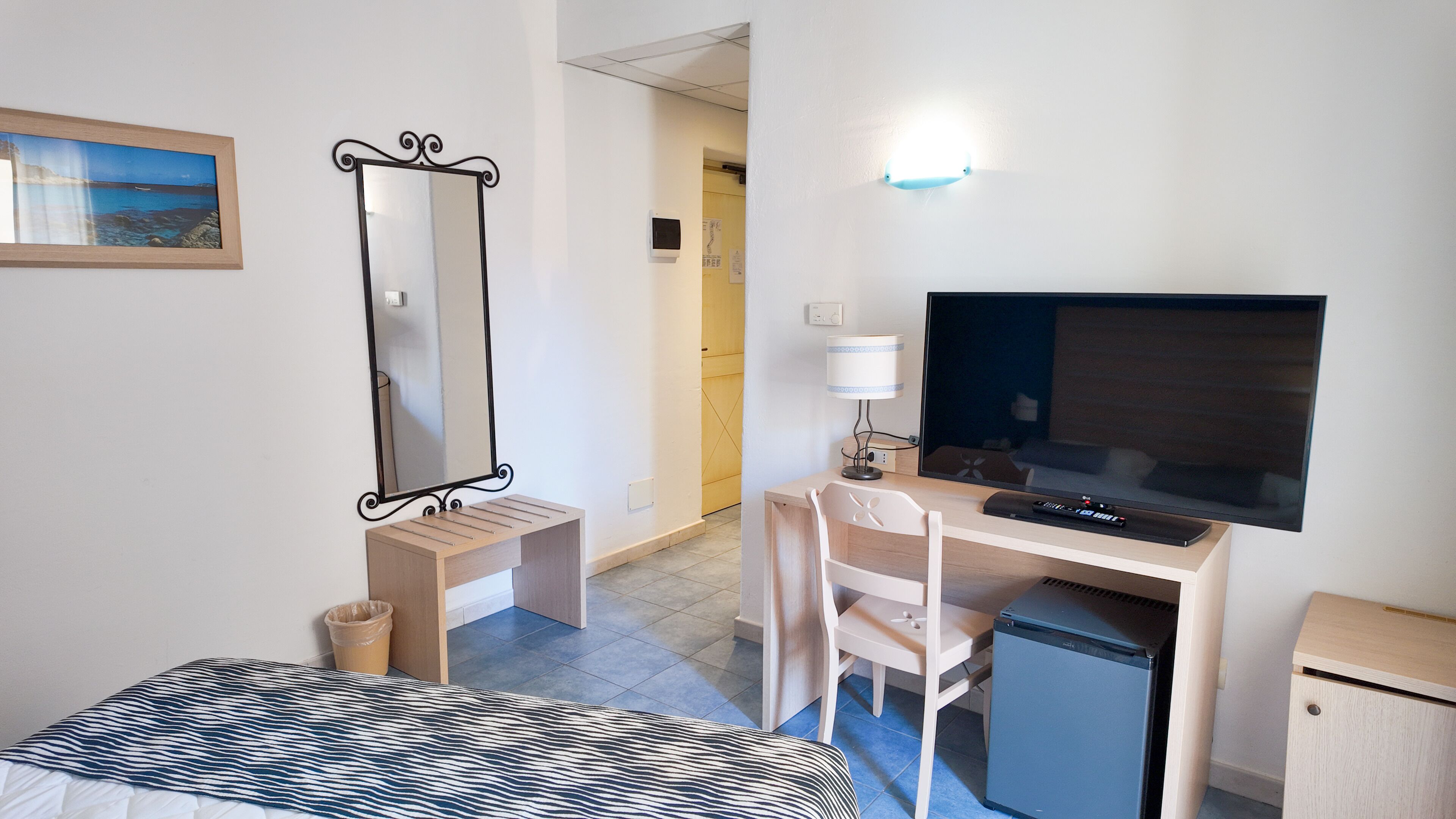 A hotel room with a bed, a desk with a computer, a TV, a small fridge, and a mirror.