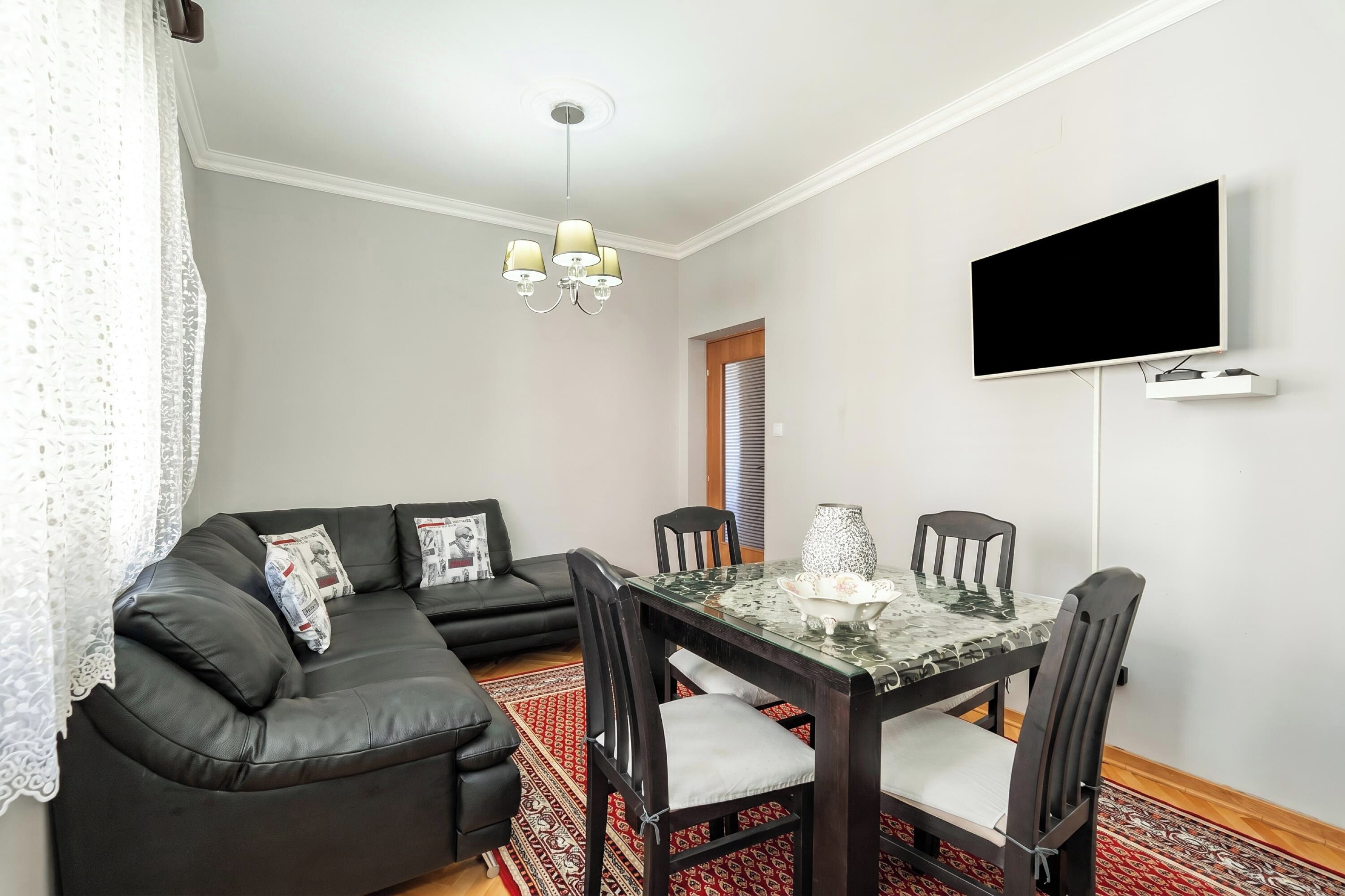Una sala de estar con un sofá de cuero negro, una mesa de comedor con cubierta de vidrio y sillas, un televisor de pantalla plana montado en la pared y una alfombra estampada.