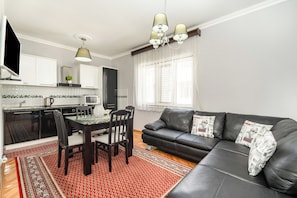 A modern kitchen with white cabinets, a dining table with chairs, a black leather sofa with patterned cushions, and a red patterned rug. - Seafront Haven Darka (Bijela)