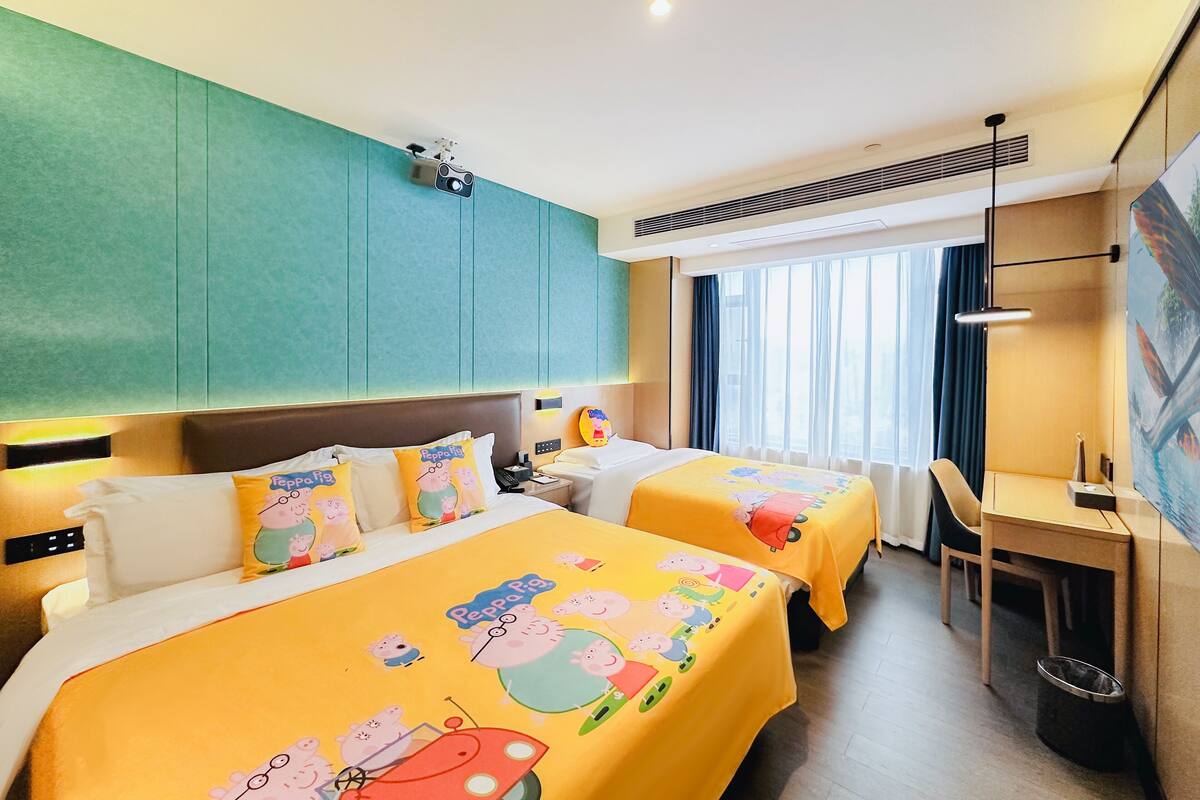 A hotel room with two beds, each with a Peppa Pig-themed bedspread, a desk with a chair, a large window with curtains, and a ceiling-mounted projector.