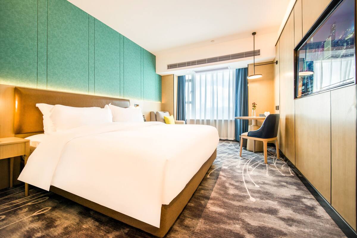 A hotel room with a large bed, a desk, a chair, and a view of the city through the window.