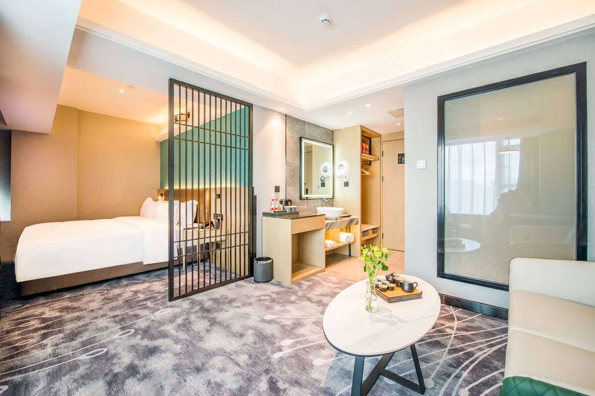 A hotel room with a bed, a small table with a vase, a bathroom with a sink and mirror, and a large window.