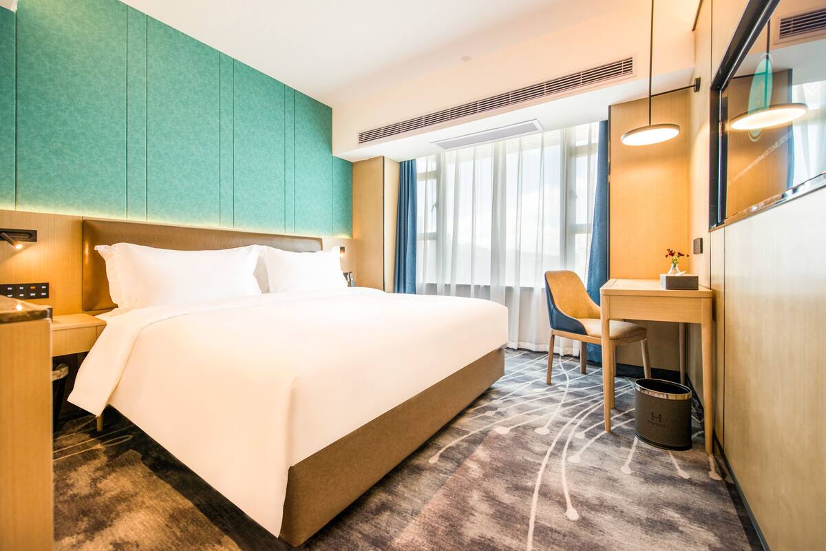 A hotel room with a bed, a desk, a chair, a trash bin, and a window with curtains.
