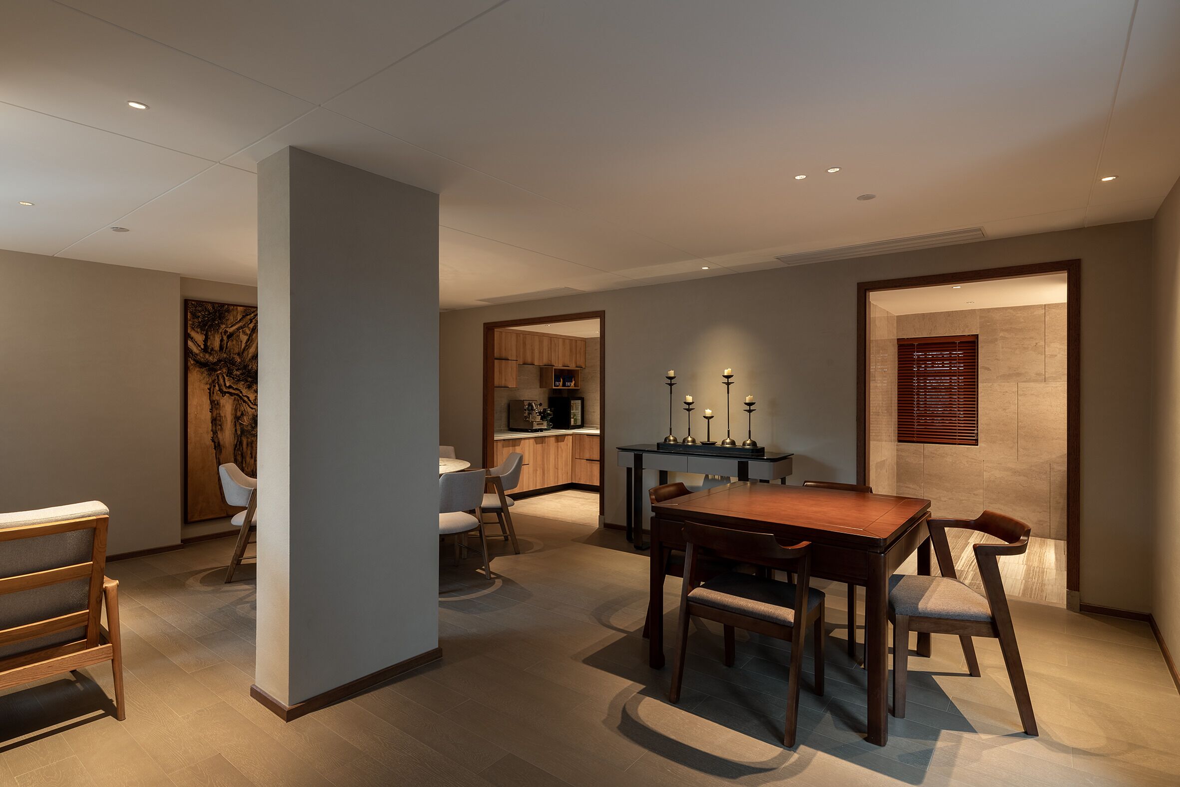 A modern dining area with a wooden table and chairs, a kitchen in the background, and a wall with a framed artwork.