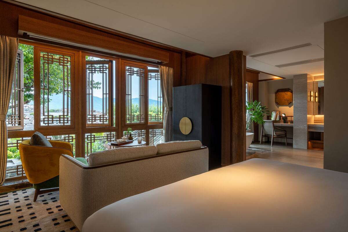 A hotel room with a large bed, a sofa, a desk, and a view of the outdoors through large windows.