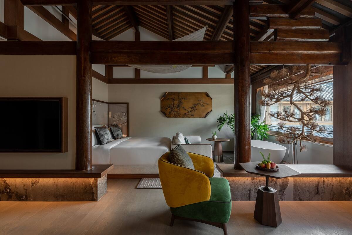 A modern interior with a bed, a yellow and green armchair, a round table with a bowl, and a large window with a view.