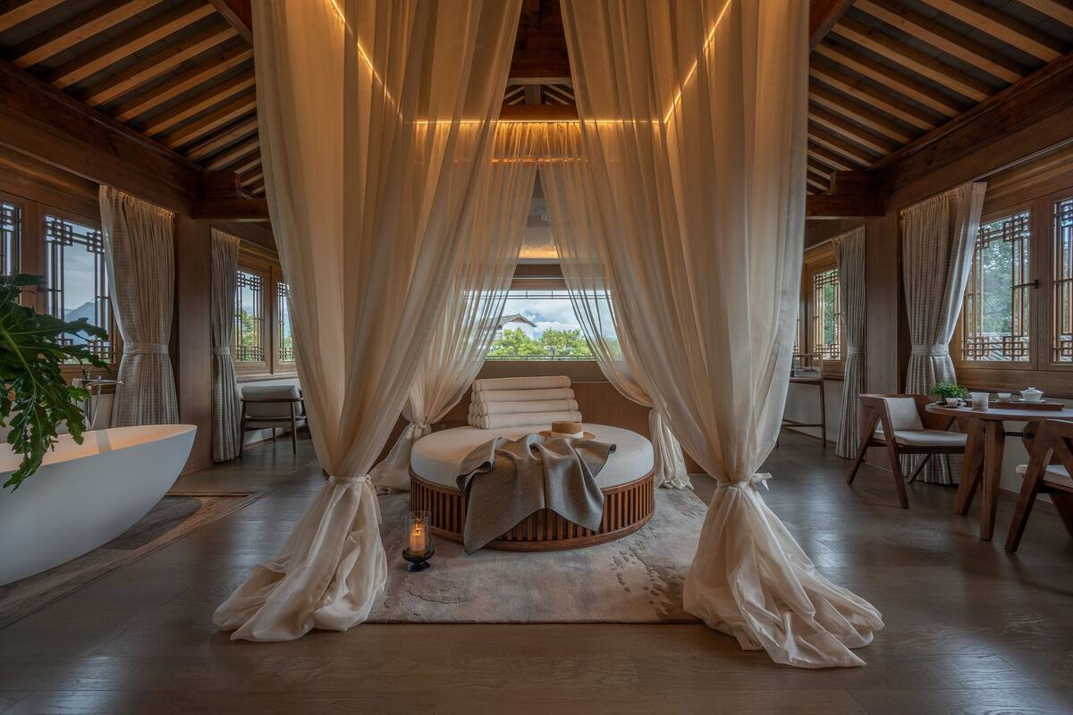 A bedroom with a circular bed, sheer curtains, a bedside table, a plant, and a view of the outdoors.