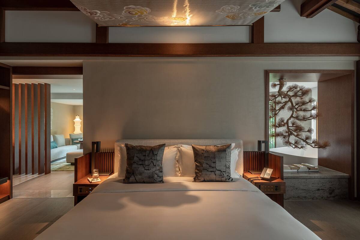 A modern hotel room with a large bed, two bedside tables with lamps, a decorative wall piece, and a view of a tree through a window.