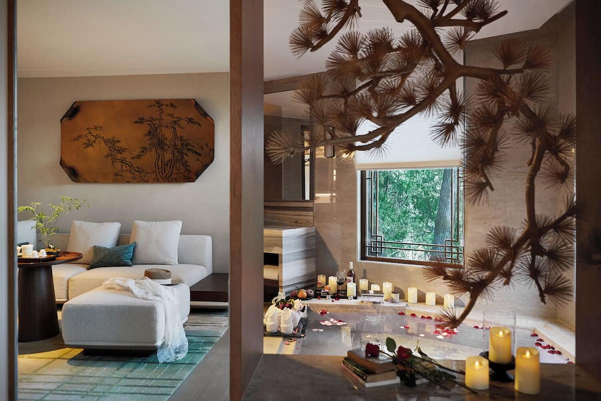 A modern bedroom with a large bed, a wall-mounted artwork, a window with a view of trees, and a decorative tree branch sculpture.