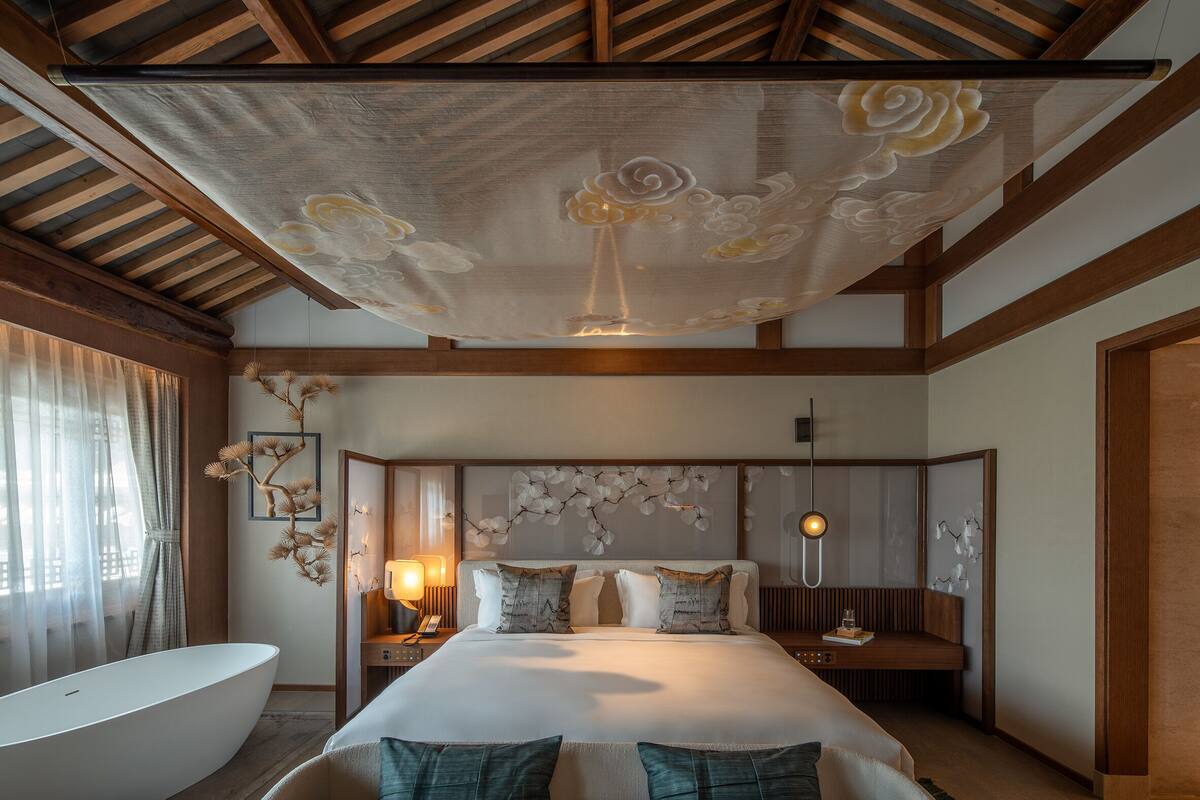 A bedroom with a large bed, a bedside table with a lamp, a wall-mounted artwork, and a ceiling with a floral design.