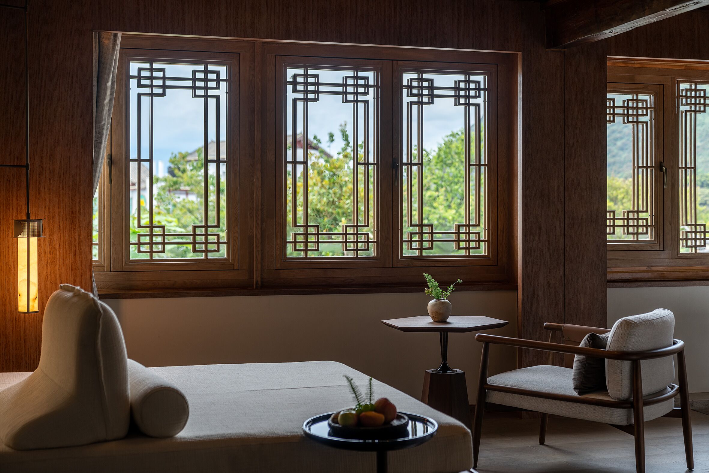 A room with a sofa, a chair, a small table with a vase and fruit, and large windows with traditional lattice designs.