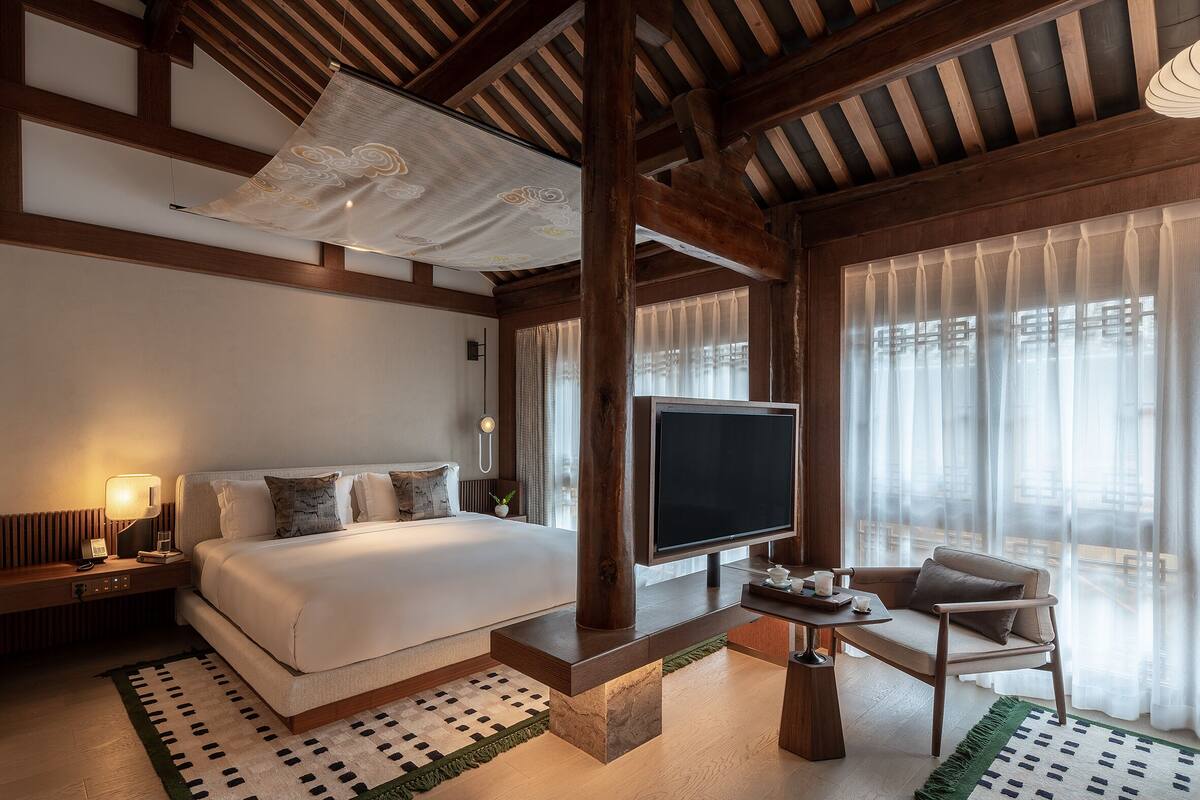 A modern hotel room with a large bed, a flat-screen TV, a seating area, and large windows.