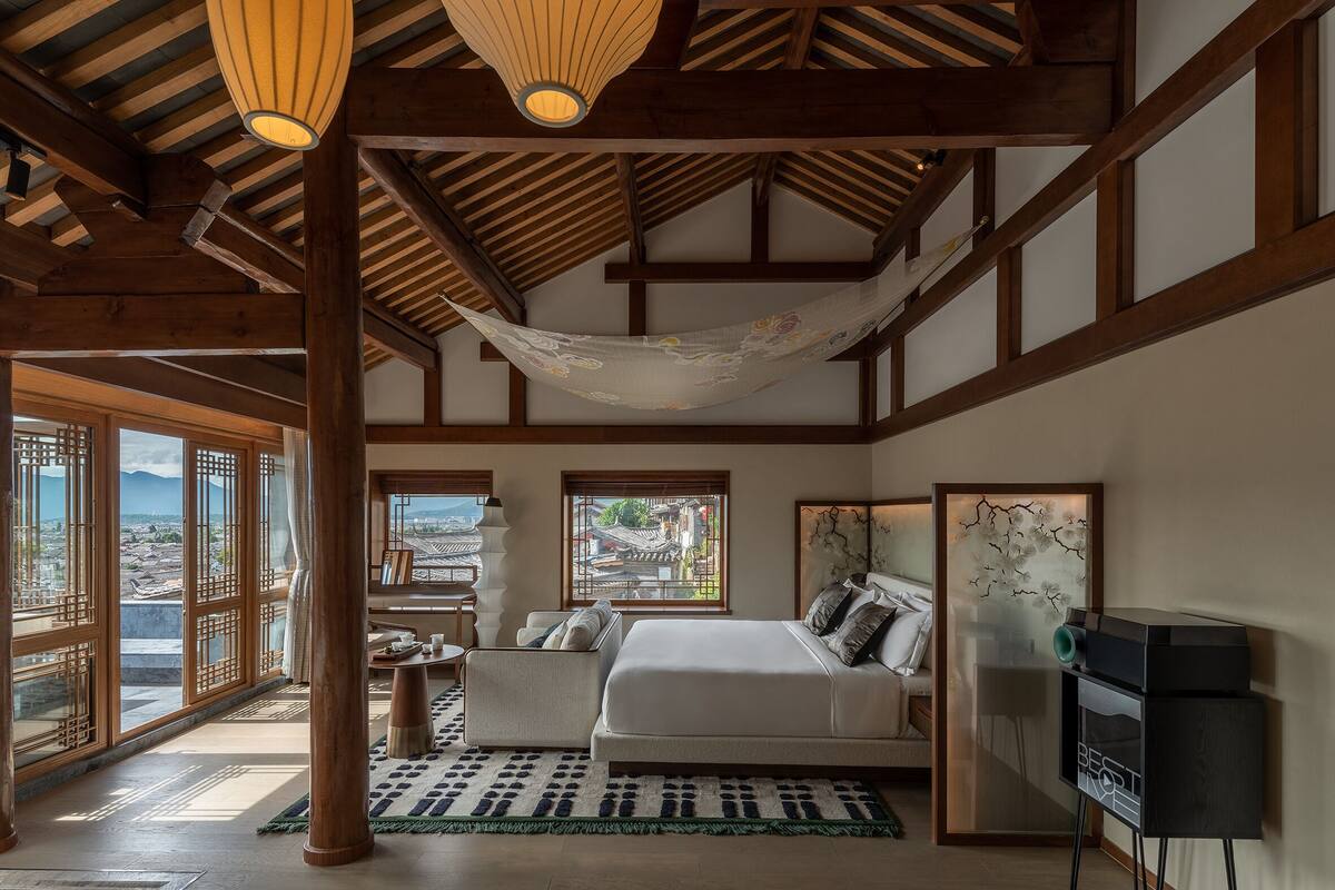 A bedroom with a large bed, a sofa, a TV, and a view of a traditional village through large windows.