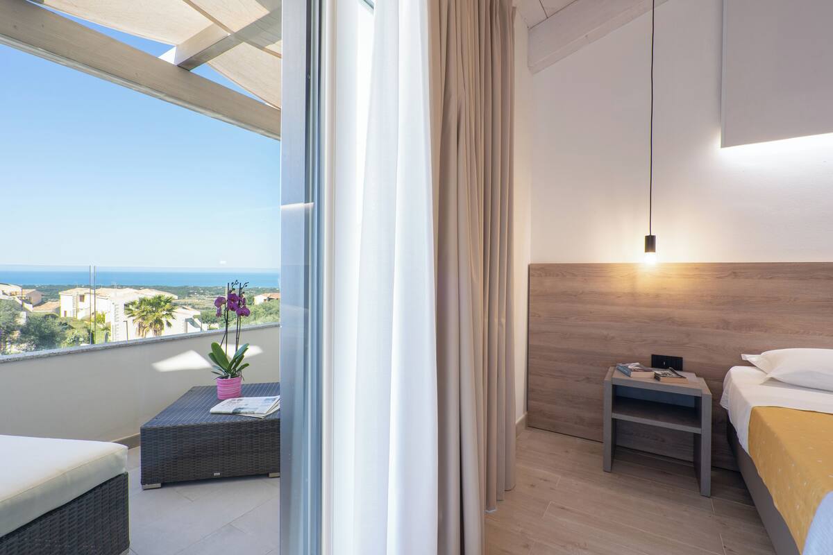 A balcony with a view of the sea, a wicker sofa, a small table with a plant, and a nightstand with a lamp.