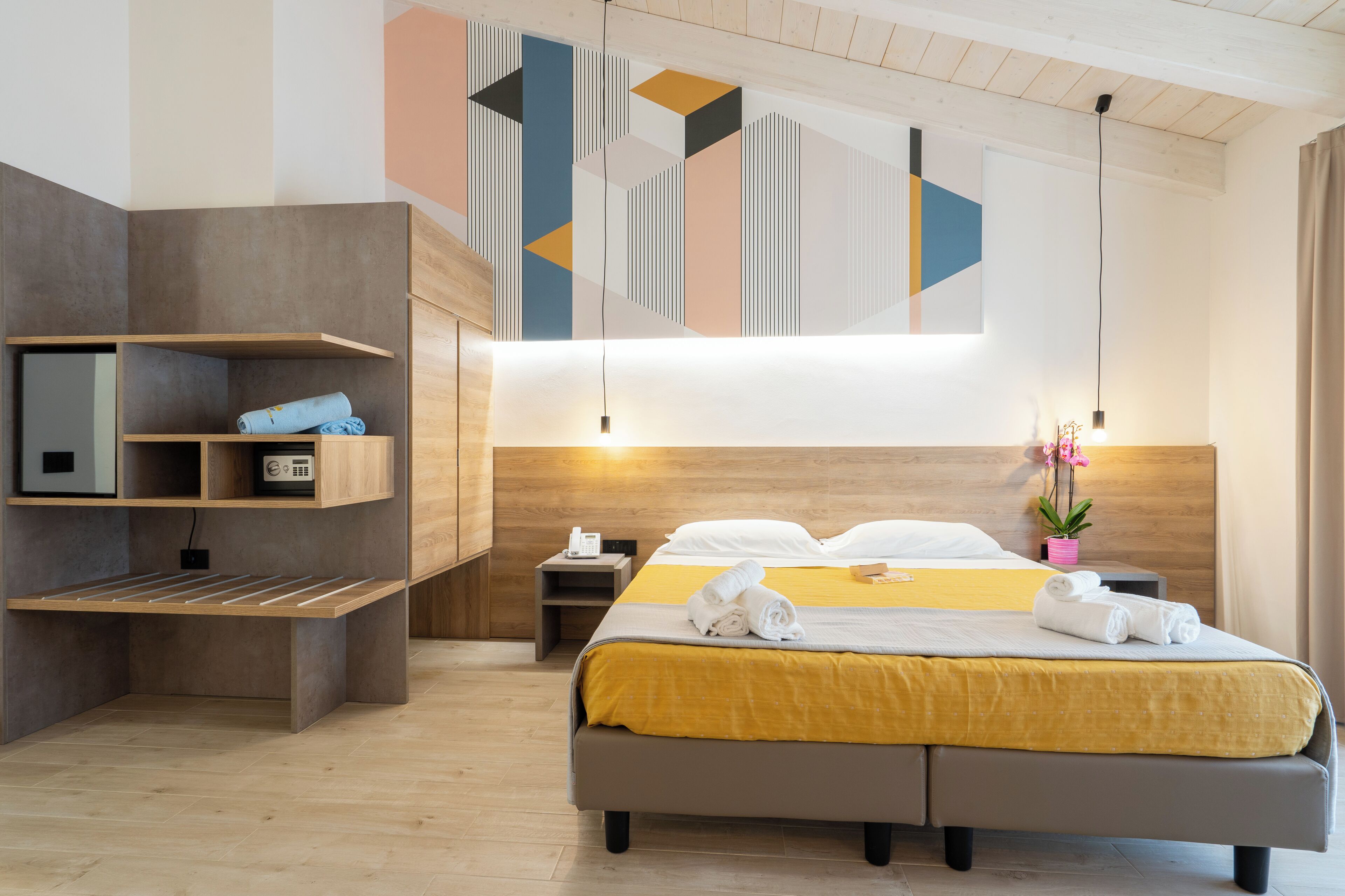 A modern hotel room with a bed, bedside tables, a built-in shelf unit, and a decorative wall panel.