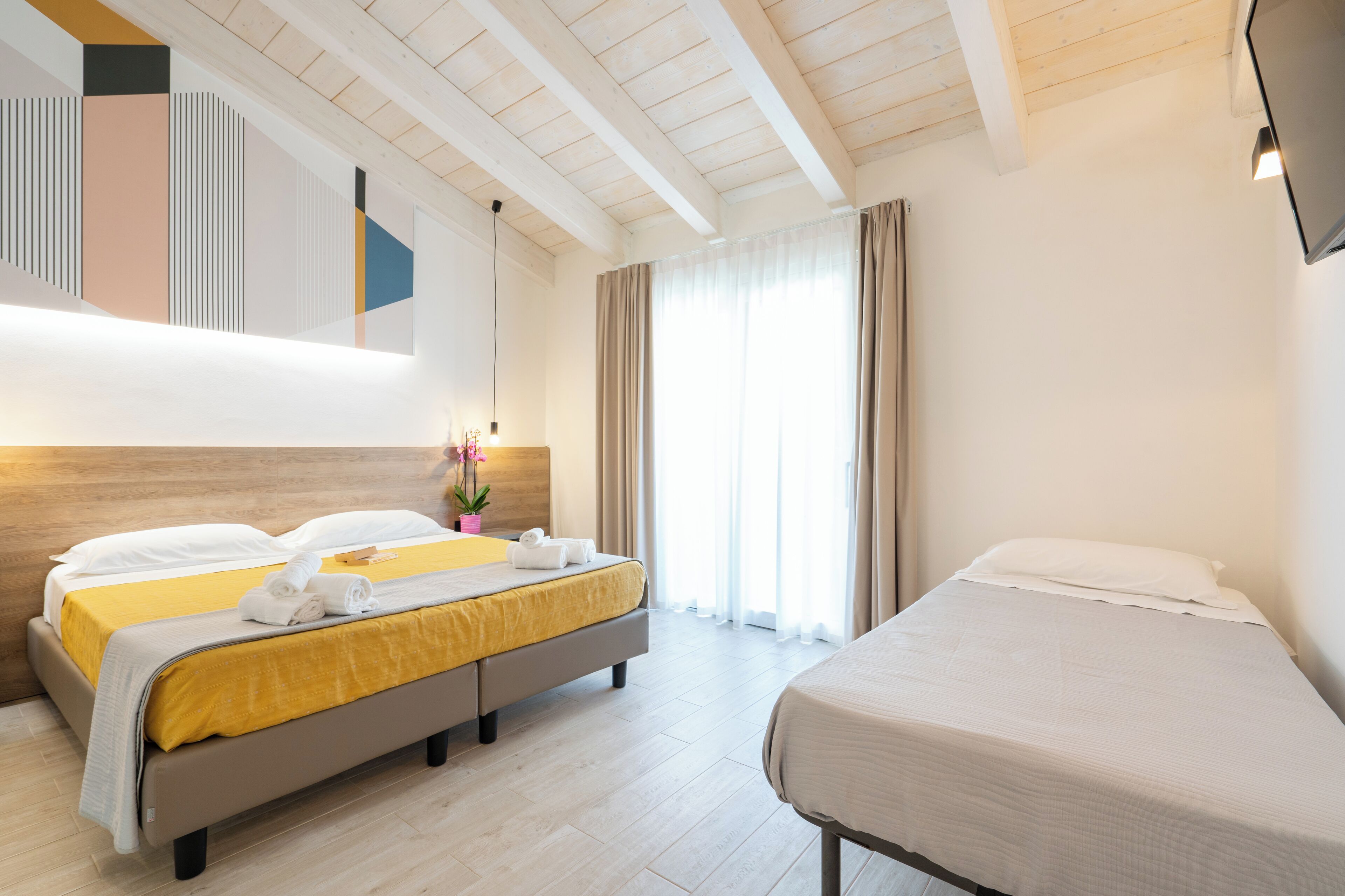 Ein Hotelzimmer mit einem großen Bett, einem kleineren Bett, einer Holzbalkendecke und einem großen Fenster mit Vorhängen.