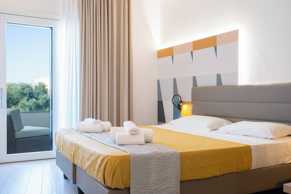A hotel room with a bed, a chair, a window, and a wall-mounted light.