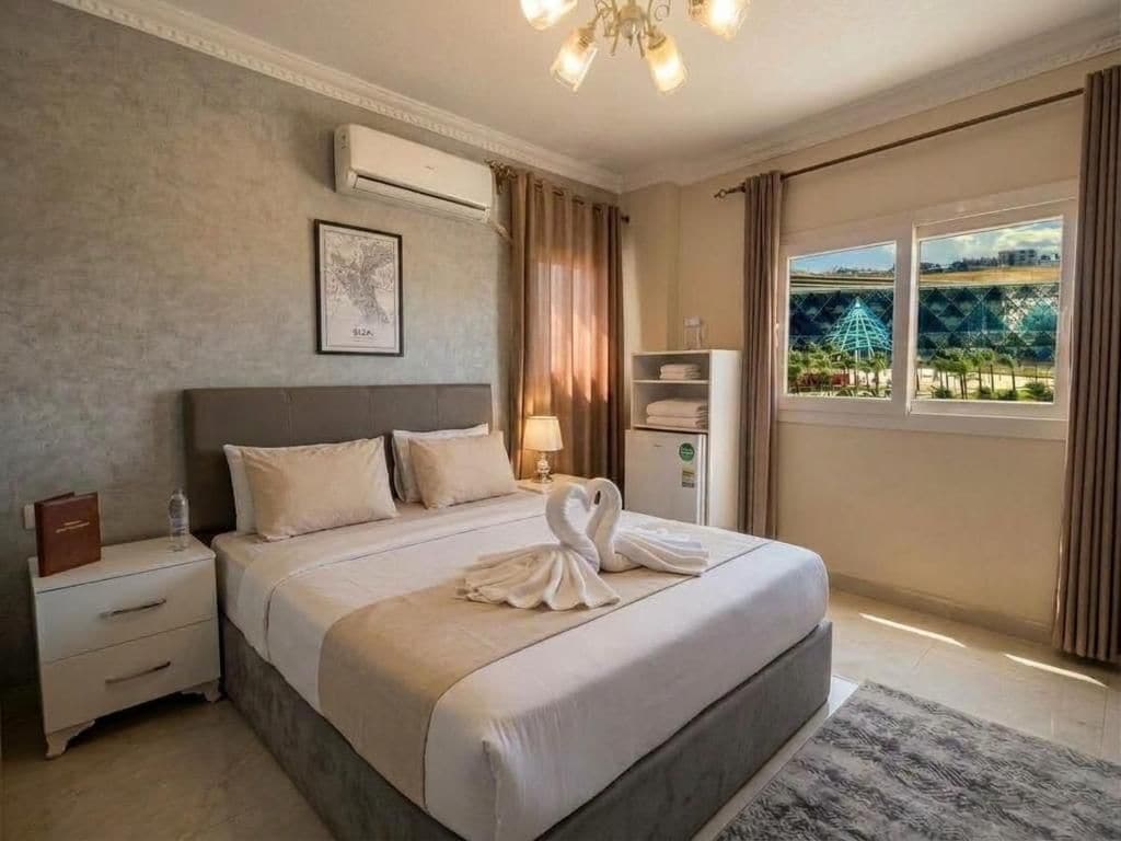 Een hotelkamer met een groot bed, een nachtkastje, een lamp, een airconditioner, een raam met uitzicht en een lijstje aan de muur.
