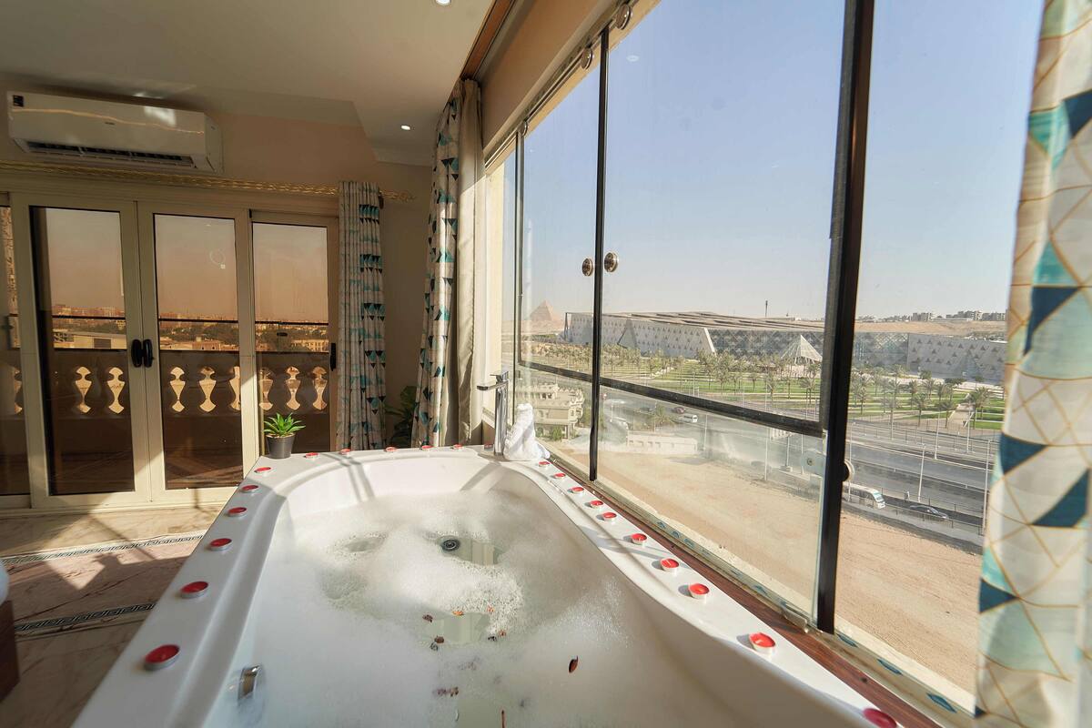 A room with a large bathtub, a view of a cityscape, and a balcony with a railing.