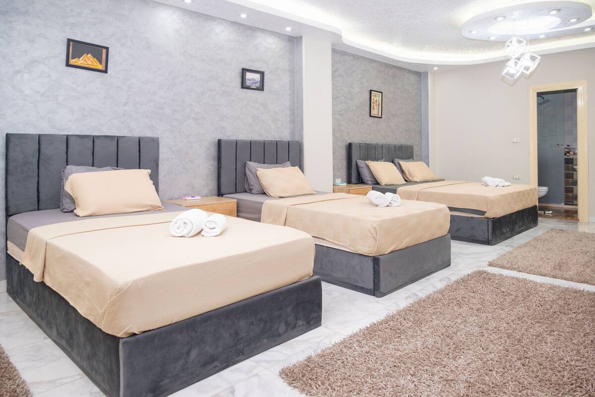A hotel room with three beds, each with a grey headboard and beige bedding. There are framed pictures on the walls and a ceiling light fixture.