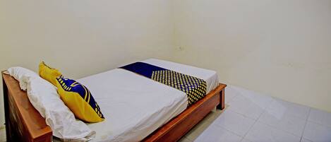Una cama individual con estructura de madera, ropa de cama blanca y dos almohadas decorativas. La habitación tiene paredes lisas y piso de baldosas.
