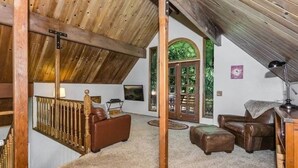 A spacious loft bedroom with a sloped ceiling, wooden beams, a large window, and a cozy seating area with a sofa and ottoman. - Lake cabin w/ big pines close to lake and village (Lake Arrowhead)