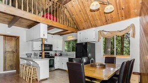 A spacious kitchen with white cabinets, a dining area with a wooden table and chairs, and a view of the outdoors through large windows. - Lake cabin w/ big pines close to lake and village (Lake Arrowhead)