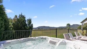 Uma banheira de hidromassagem com cadeiras de lounge, cercada por uma grade metálica, com vista para um campo gramado e montanhas ao fundo.