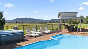 Piscina com espreguiçadeiras e uma banheira de hidromassagem, cercada por uma grade e um pergolado, com vista para colinas distantes.
