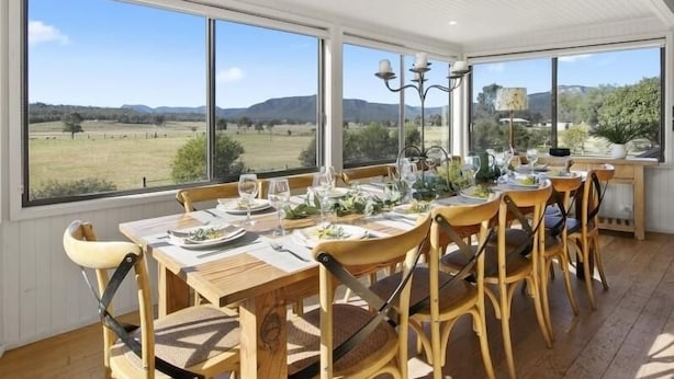 Uma área de refeições com mesa posta para seis pessoas, grandes janelas com vista para uma paisagem rural e um lustre sobre a mesa.