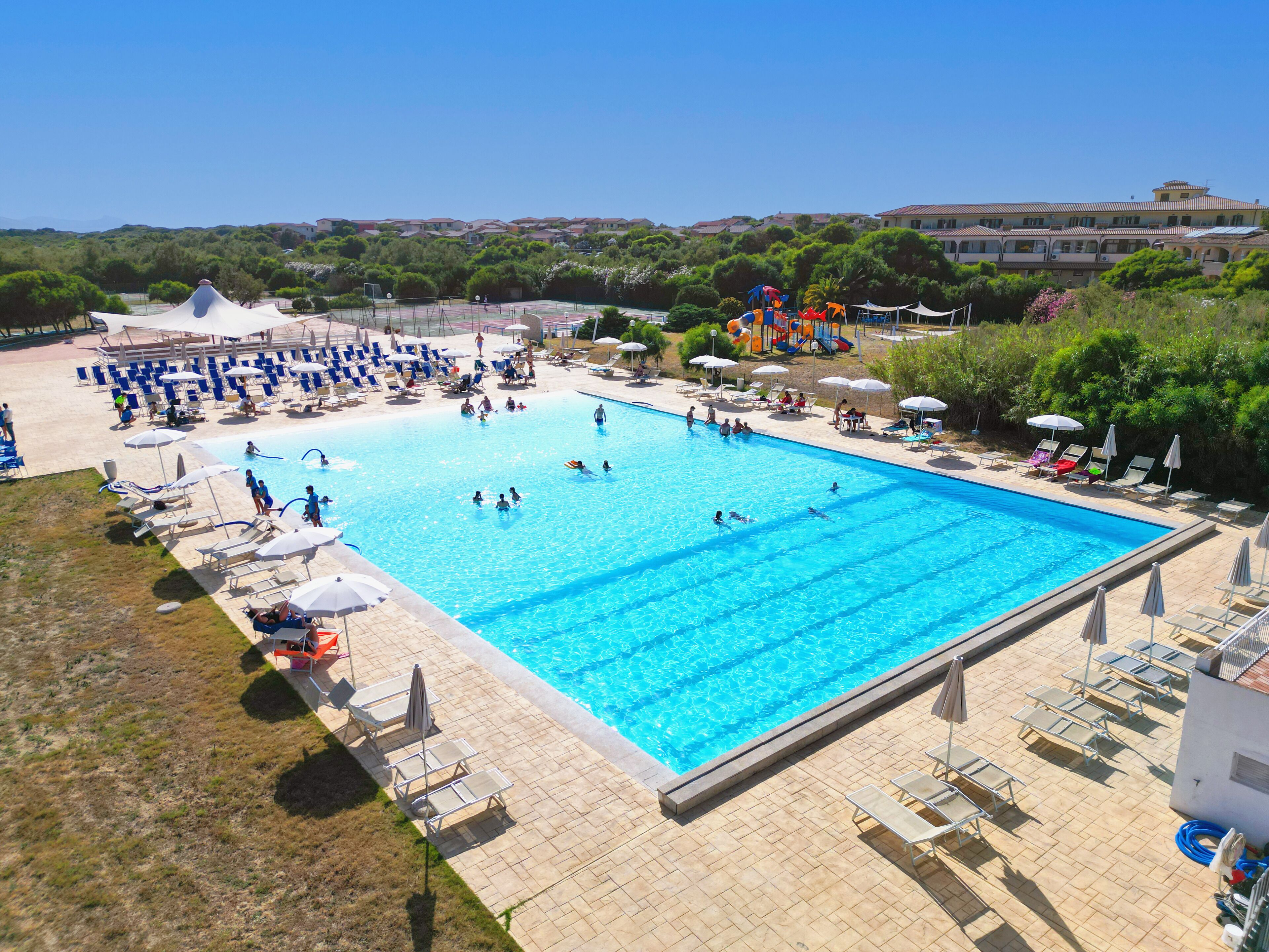 Uma piscina retangular grande, rodeada por espreguiçadeiras e guarda-sóis, com vista para uma área residencial ao fundo.