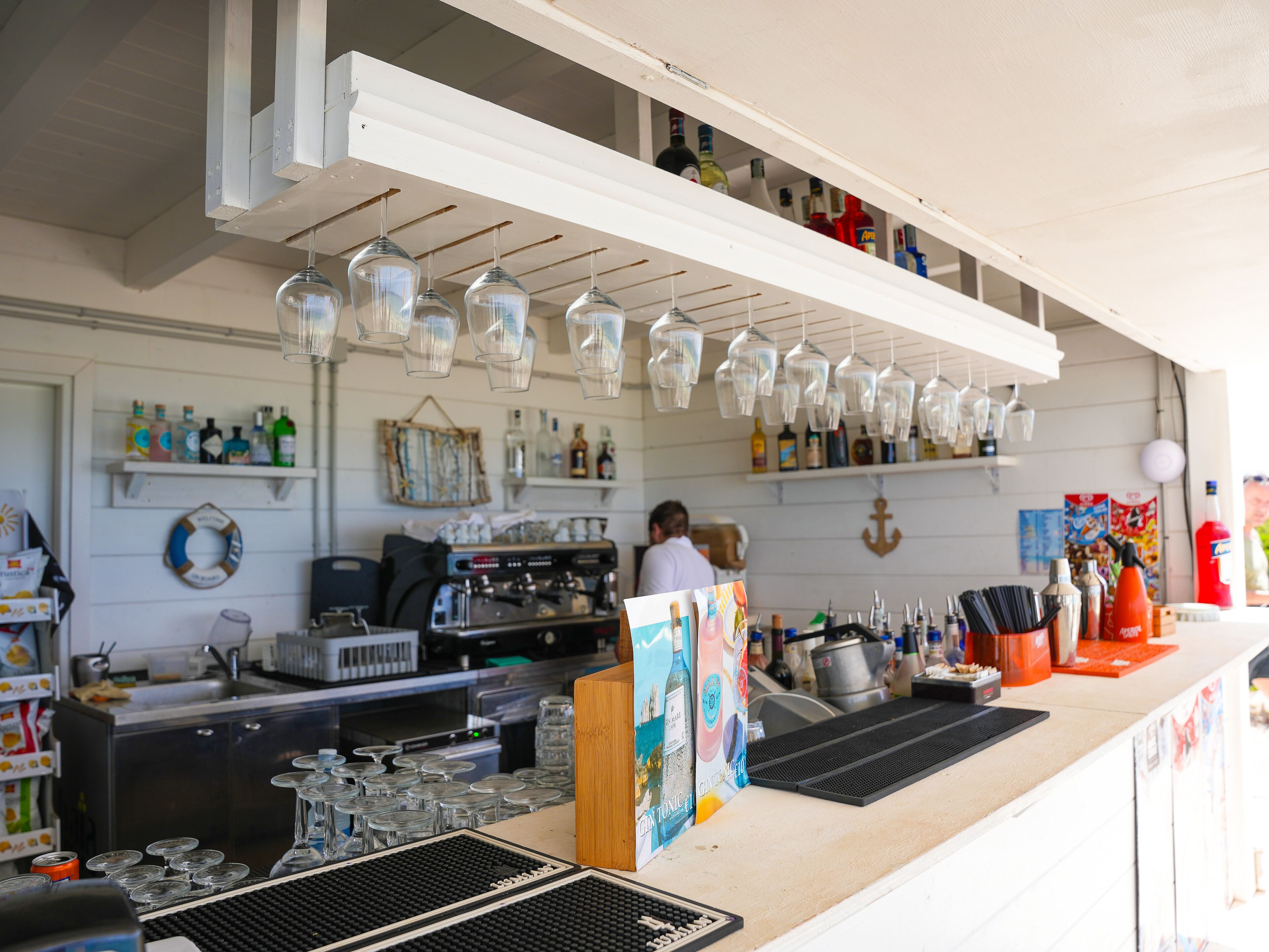 A bar area with a coffee machine, shelves with bottles, and hanging glassware.