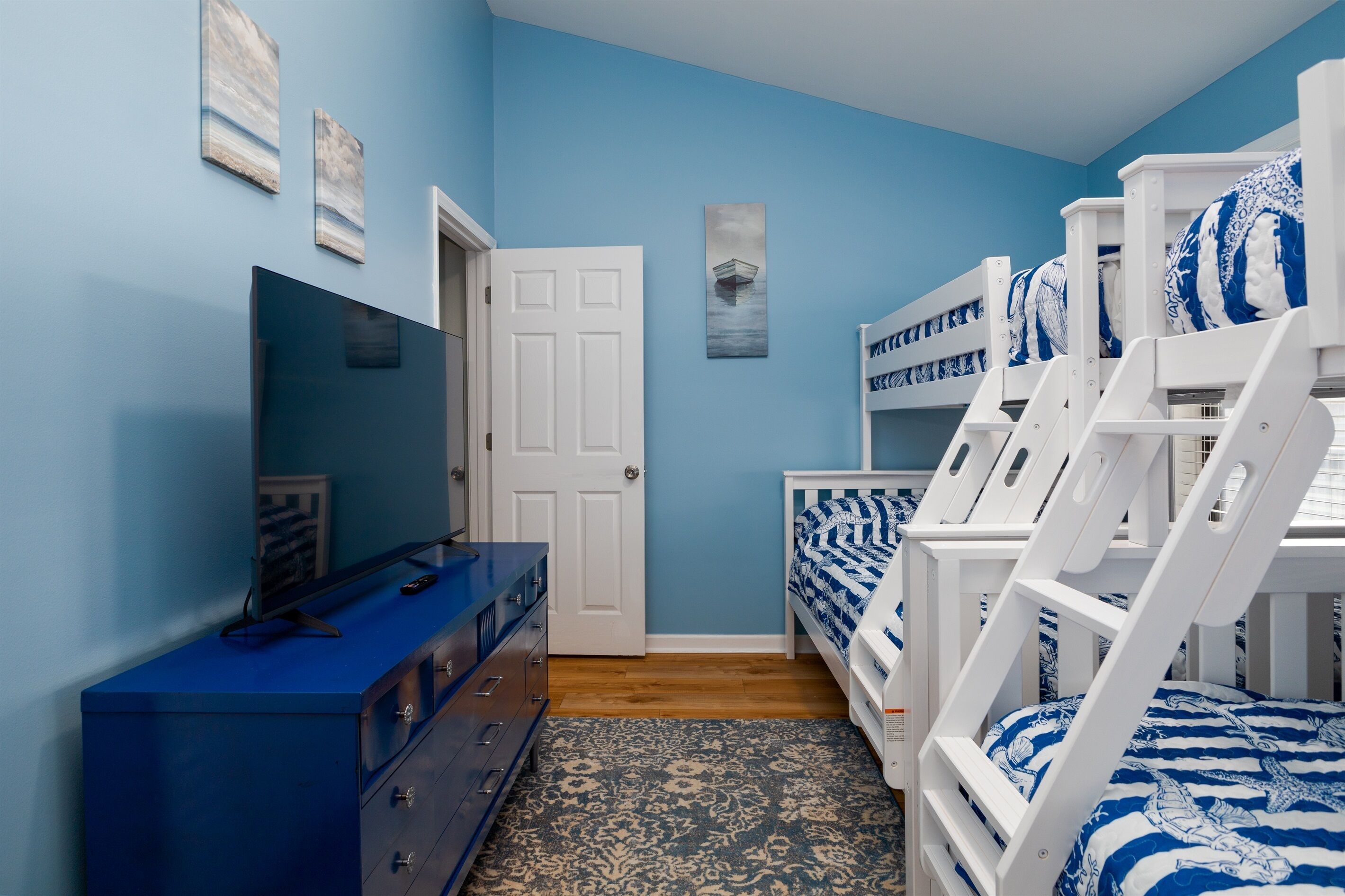 A bedroom with blue walls, a white door, a dresser, bunk beds, and a patterned rug.